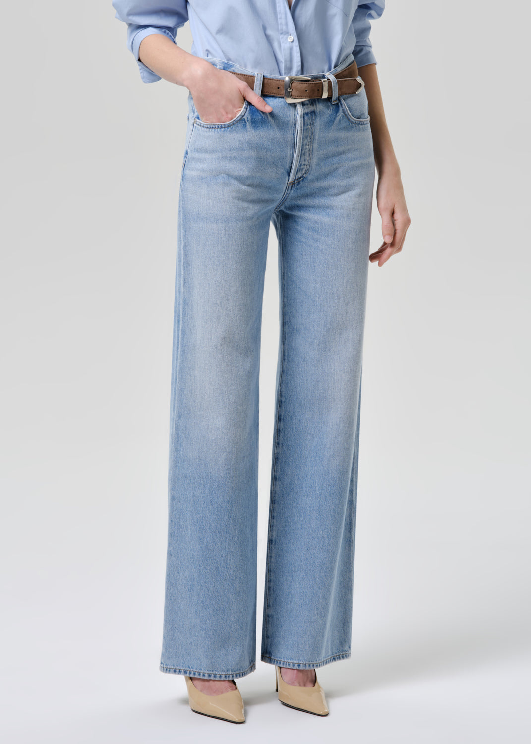 A model wearing light-wash blue wide-leg denim jeans with a brown leather belt, styled with a light blue shirt and beige pointed-toe heels against a grey background.