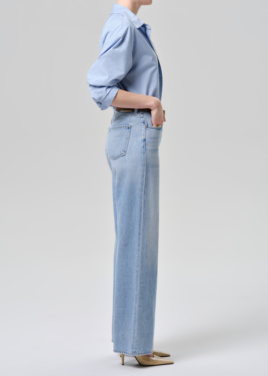 A side-profile studio shot of a model wearing light-wash blue wide-leg denim jeans and a matching light blue button-down shirt, paired with beige pointed-toe heels, against a grey background.