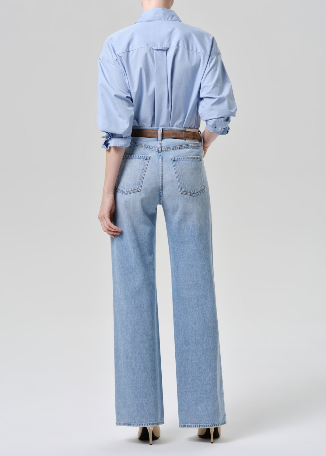 A model shown from the back wearing a light blue button-up shirt and matching wide-leg light blue jeans, styled with a brown belt and black heels against a grey background.