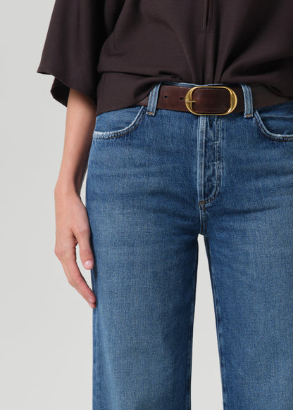 Close-up of a person wearing blue jeans with a brown leather belt featuring a gold-tone oval buckle, paired with a dark brown top against a grey background.