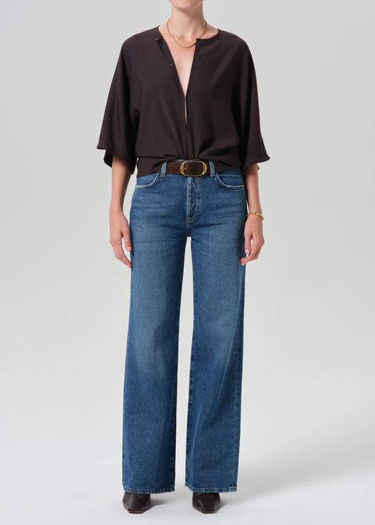 A model wearing a dark brown short-sleeve blouse tucked into high-waisted, wide-leg blue jeans with a brown belt, standing against a grey background.