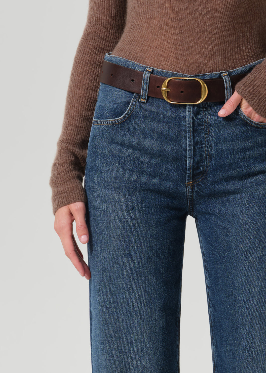 Close-up of a person wearing blue denim jeans with a brown leather belt featuring a gold-tone oval buckle, paired with a brown ribbed knit sweater against a grey background.