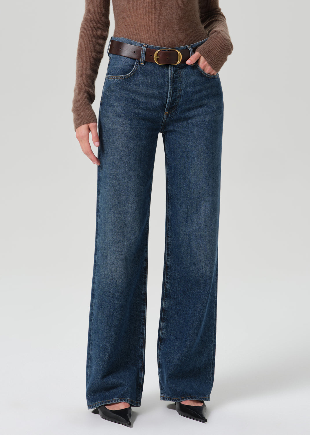 A model wearing a pair of dark blue wide-leg jeans with a brown belt, styled with a brown knit top and black pointed-toe heels against a grey background.