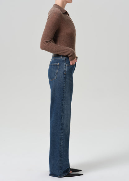 A side-profile studio shot of a model wearing a brown long-sleeve knit polo shirt and high-waisted, wide-leg blue denim jeans against a grey background.