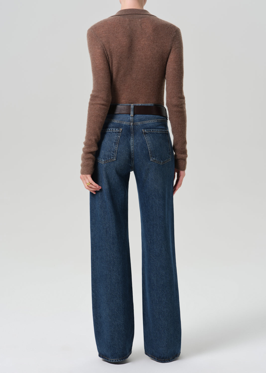 A model shown from the back wearing a brown long-sleeve knit top and high-waisted, wide-leg blue jeans with a dark brown belt, against a grey background.