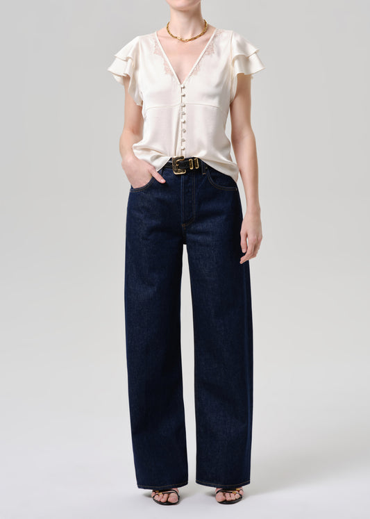 A model wearing a cream ruffled short-sleeve blouse and dark navy wide-leg jeans with a gold belt, standing against a grey background.