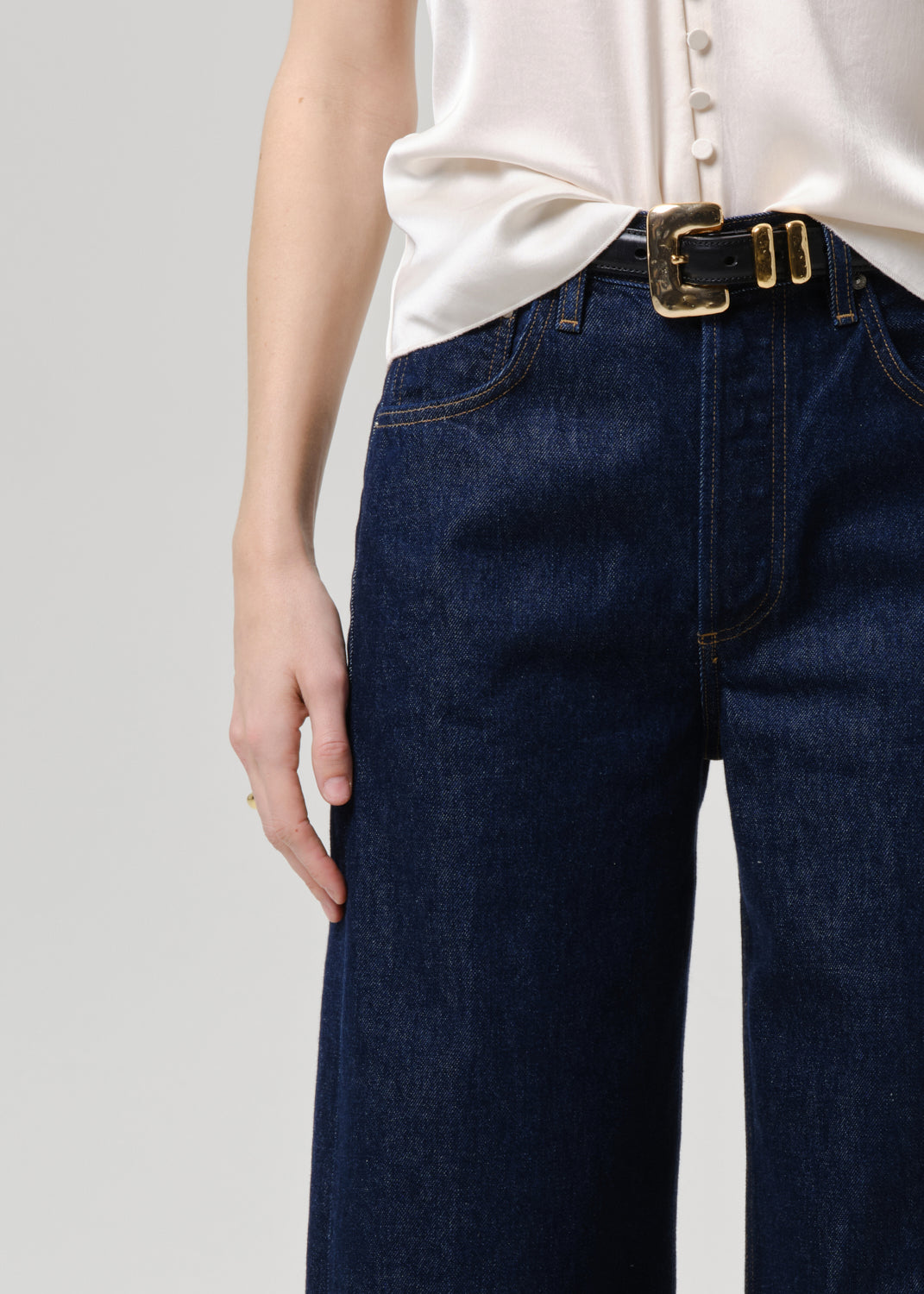A close-up of a person wearing dark blue wide-leg denim jeans with a black belt featuring a gold buckle, paired with a cream-colored blouse.