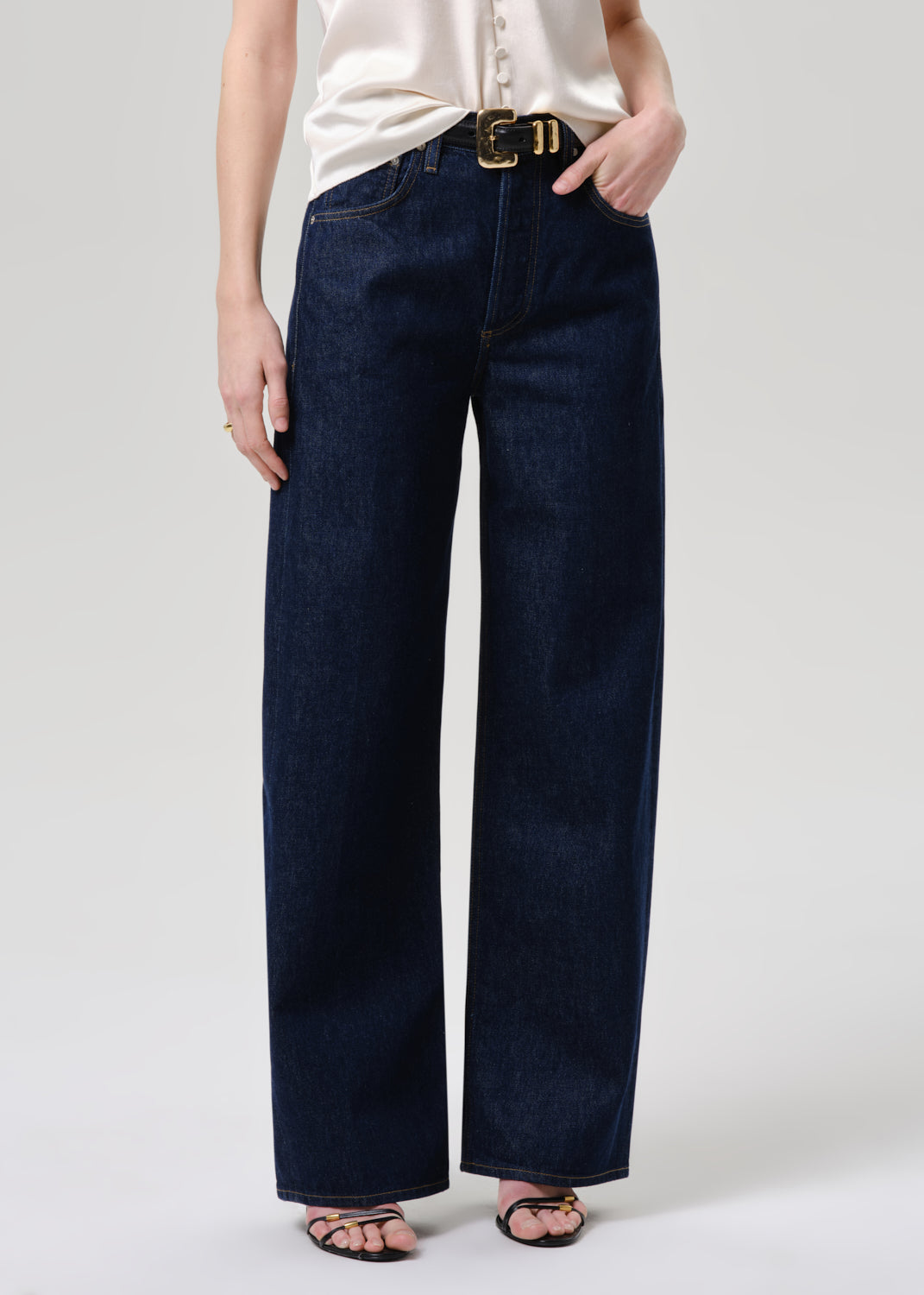A person wearing dark indigo wide-leg jeans with a gold-tone belt buckle, paired with a cream blouse and black sandals, shown from the waist down against a grey background.