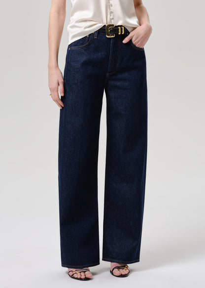 A person wearing dark indigo wide-leg jeans with a gold-tone belt buckle, paired with a cream blouse and black sandals, shown from the waist down against a grey background.