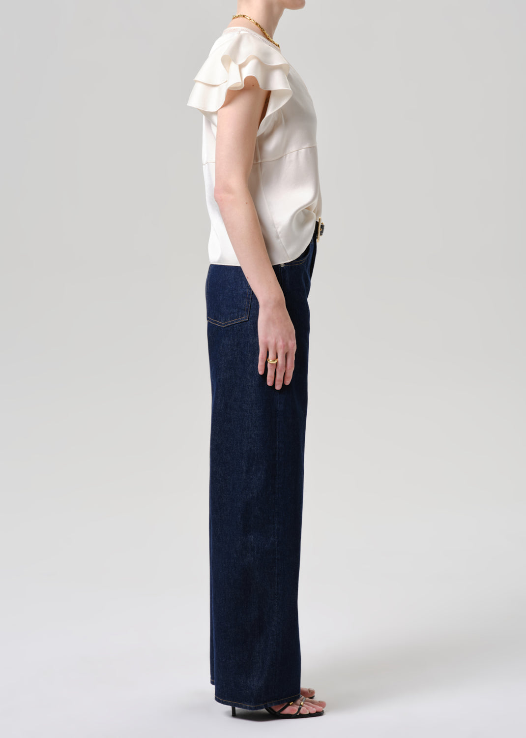 A side-profile view of a model wearing a cream ruffled short-sleeve blouse and dark navy wide-leg denim pants, paired with black sandals, against a grey background.