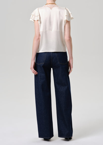 A model shown from the back wearing a cream-colored short-sleeve blouse with ruffled details and dark blue wide-leg denim jeans, set against a grey background.
