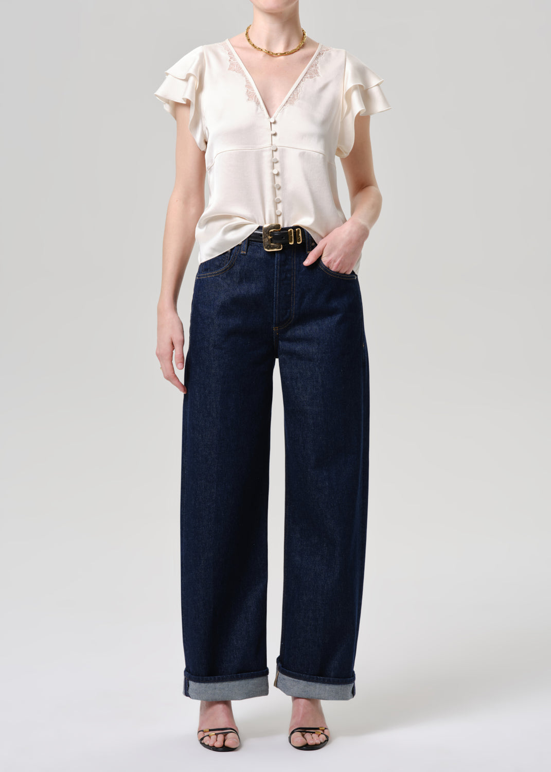 A model wearing a cream-colored ruffled short-sleeve blouse and dark blue wide-leg denim jeans with rolled cuffs, styled with a gold belt and black sandals against a grey background.