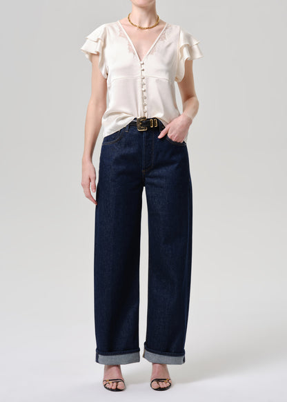 A model wearing a cream-colored ruffled short-sleeve blouse and dark blue wide-leg denim jeans with rolled cuffs, styled with a gold belt and black sandals against a grey background.