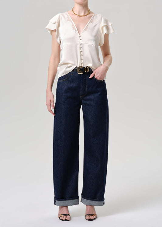 A model wearing a cream-colored ruffled short-sleeve blouse and dark blue wide-leg denim jeans with rolled cuffs, styled with a gold belt and black sandals against a grey background.