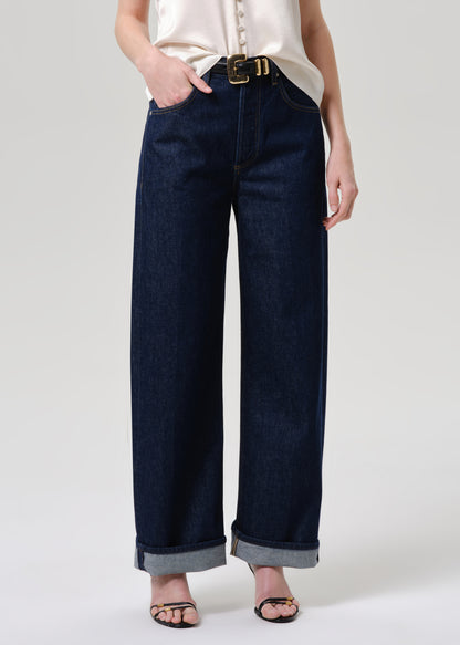 A model wearing dark blue wide-leg denim jeans with a rolled hem, paired with a cream blouse and black sandals, shown from the waist down on a grey background.