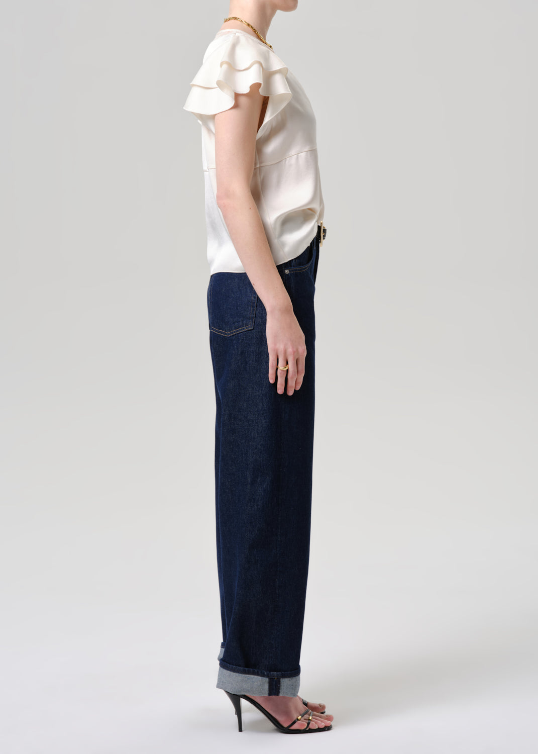 A model shown from the side wearing a cream ruffled short-sleeve blouse, dark blue wide-leg jeans with rolled cuffs, and black high-heeled sandals against a grey background.