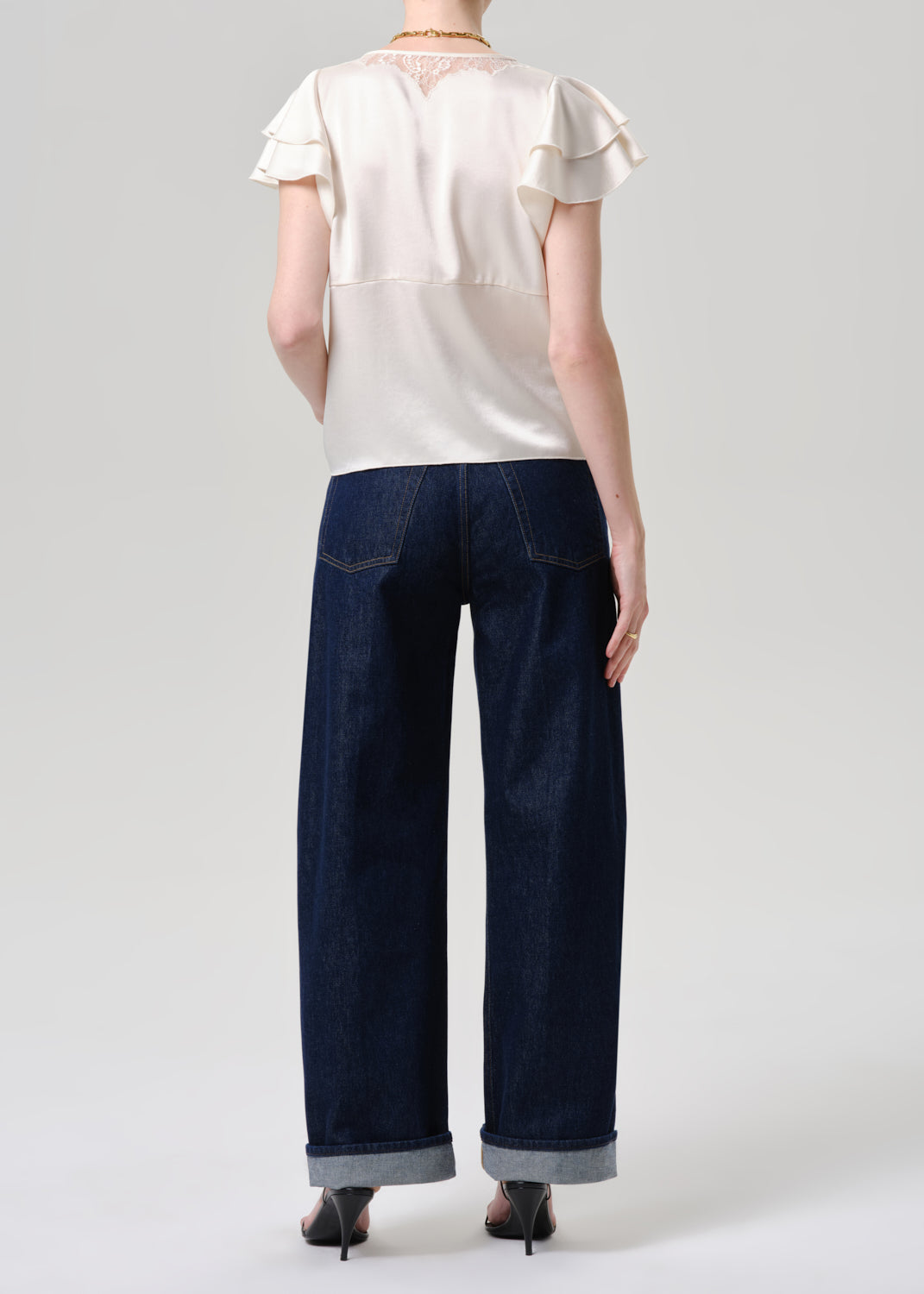 A model shown from the back wearing a cream ruffled short-sleeve blouse, dark blue wide-leg jeans with rolled cuffs, and black high-heeled sandals against a grey background.