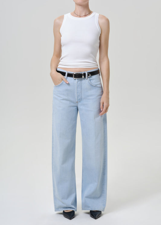 Person wearing light blue wide leg jeans with a white sleeveless tank top, black belt, and black pointed-toe heels, front full body view on a grey background.