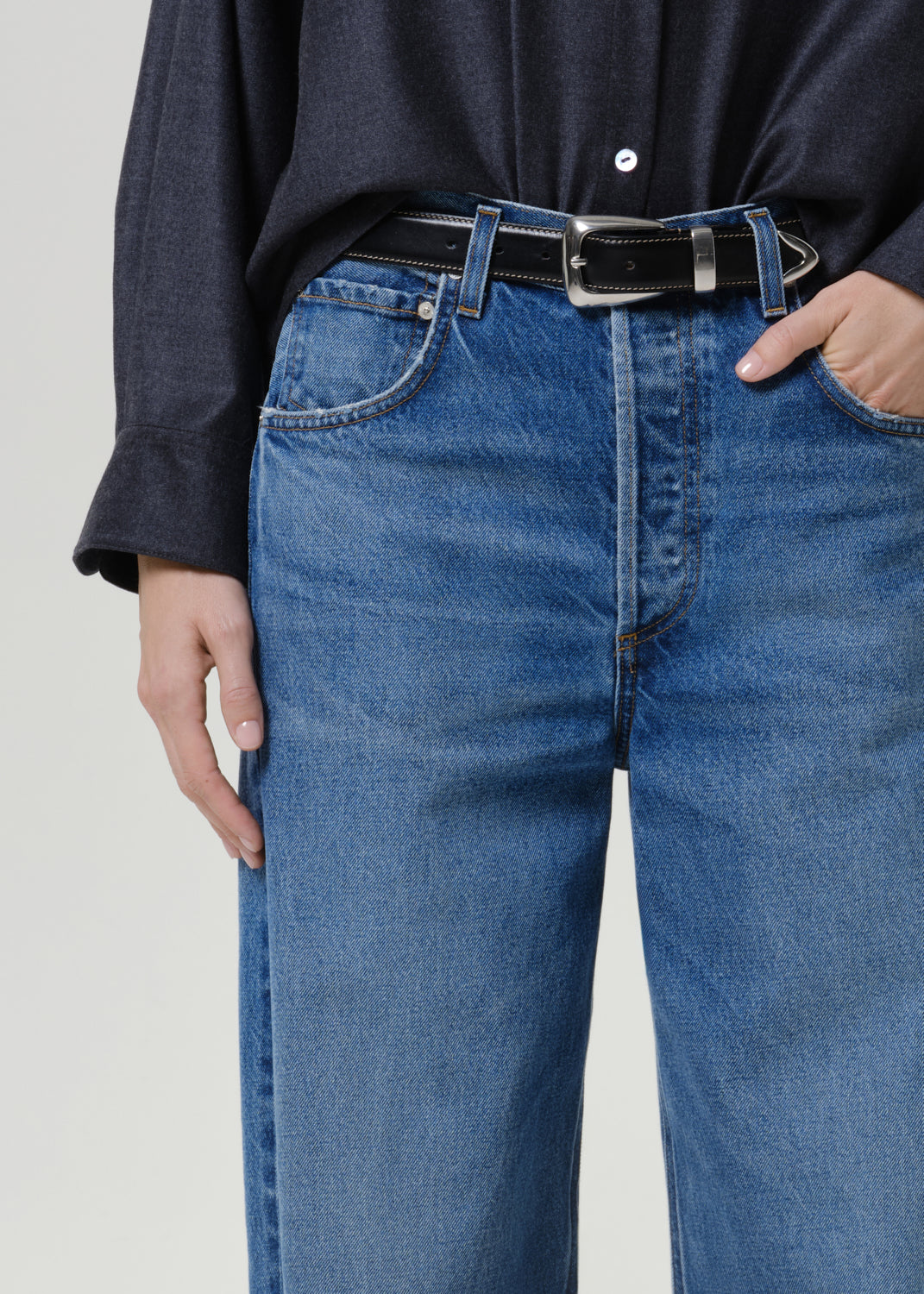 Close-up of a person wearing blue denim jeans with a black belt and a dark navy shirt, shown from the chest down against a light gray studio background.