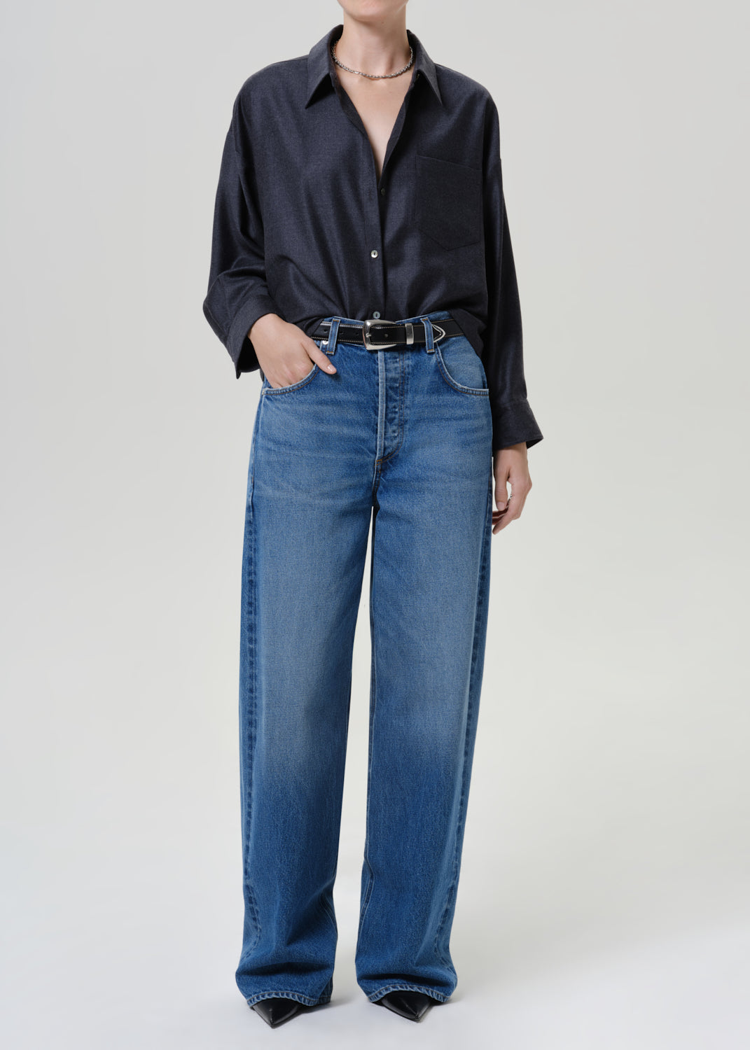 A model wearing a dark navy long-sleeve button-up shirt and high-waisted, wide-leg blue jeans with a black belt, photographed against a neutral light gray studio background.