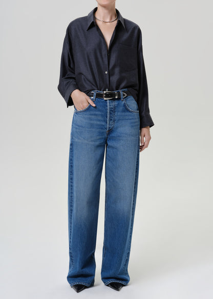 A model wearing a dark navy long-sleeve button-up shirt and high-waisted, wide-leg blue jeans with a black belt, photographed against a neutral light gray studio background.