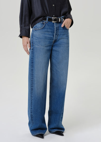 A person wearing blue wide-leg denim jeans with a black belt, paired with a dark navy button-up shirt and black pointed shoes, photographed against a light gray studio background.