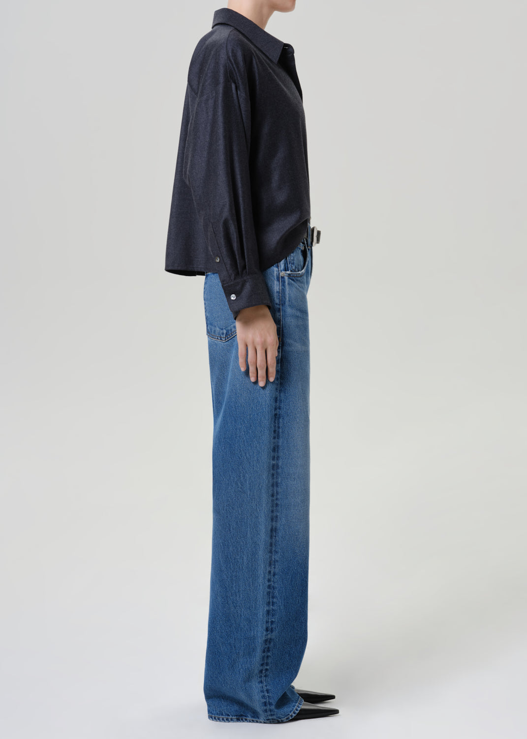 A side-profile studio shot of a model wearing a dark navy long-sleeve button-up shirt and wide-leg blue denim jeans against a light gray background.
