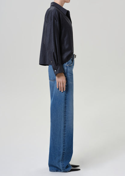 A side-profile studio shot of a model wearing a dark navy long-sleeve button-up shirt and wide-leg blue denim jeans against a light gray background.
