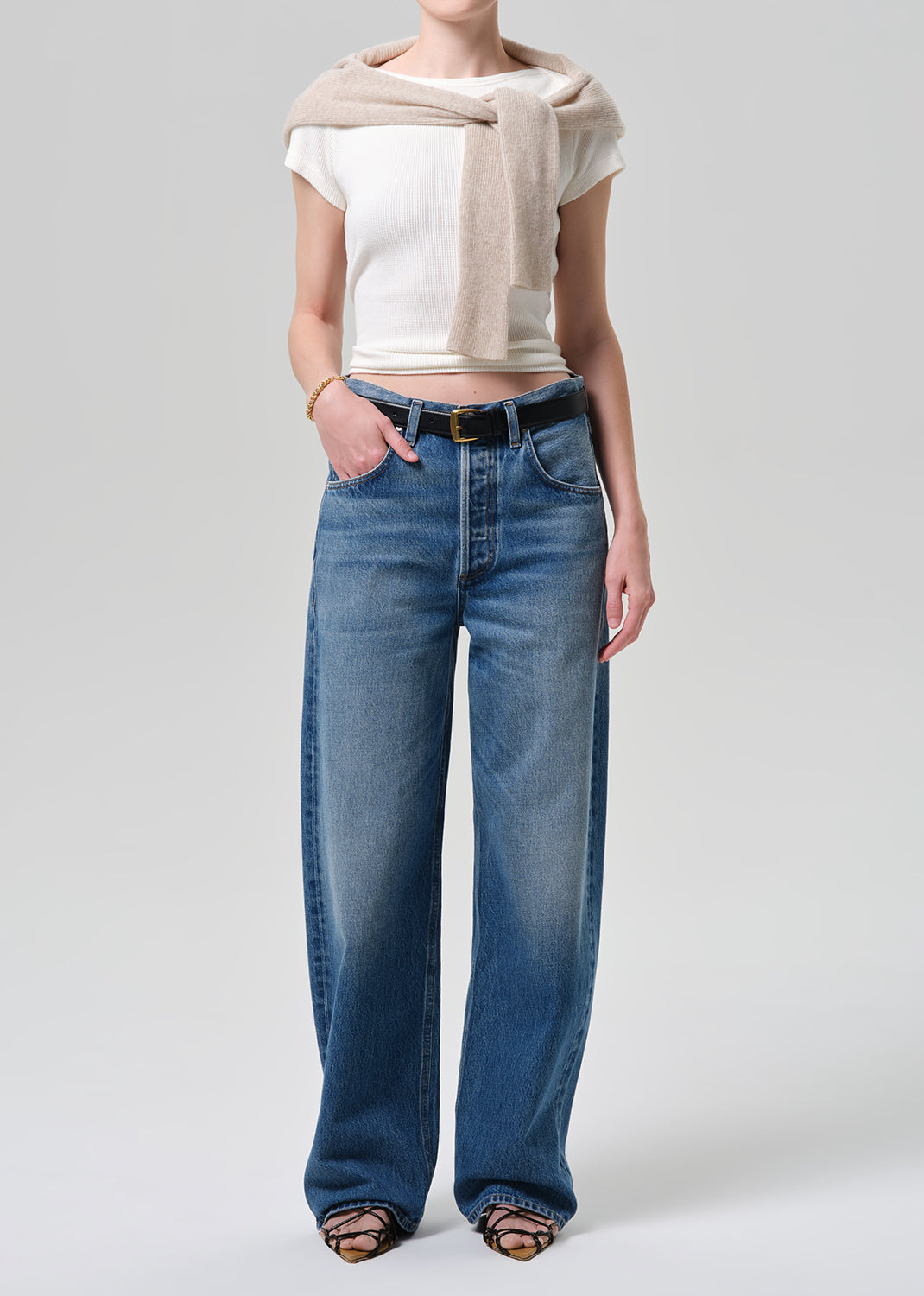 Person wearing a white crop top, beige cardigan, and blue jeans on a plain background