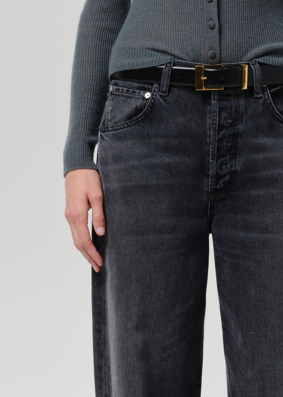 Close-up of a person wearing black washed denim jeans with a black belt featuring a gold buckle, paired with a dark ribbed top against a grey background.