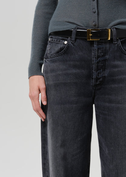 Close-up of a person wearing black washed denim jeans with a black belt featuring a gold buckle, paired with a dark ribbed top against a grey background.