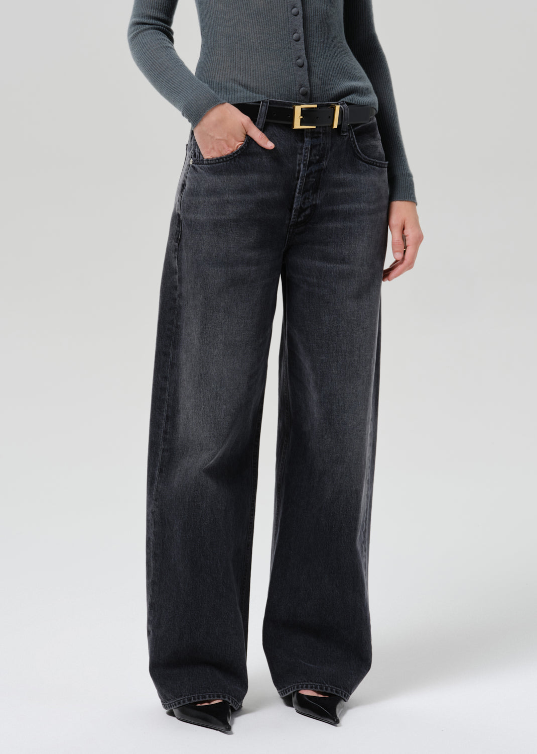 A model wearing a pair of wide-leg black denim jeans with a gold-tone belt, paired with a grey long-sleeve top and black pointed-toe heels, photographed against a grey background.