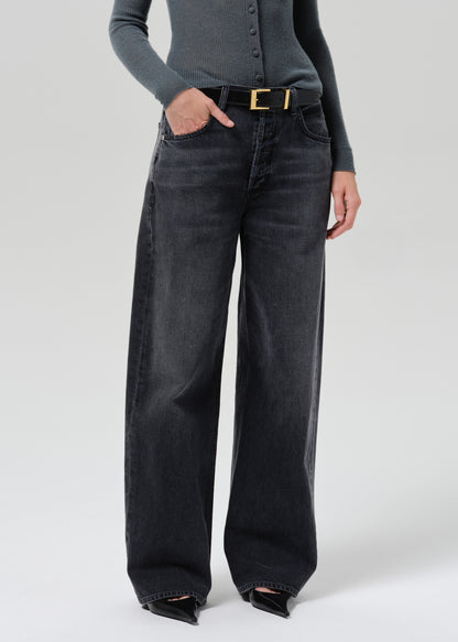 A model wearing a pair of wide-leg black denim jeans with a gold-tone belt, paired with a grey long-sleeve top and black pointed-toe heels, photographed against a grey background.