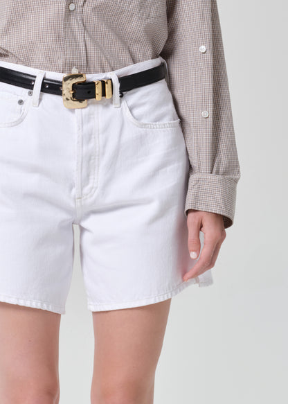 Person wearing white denim shorts with a black belt and beige checkered shirt on a light gray background