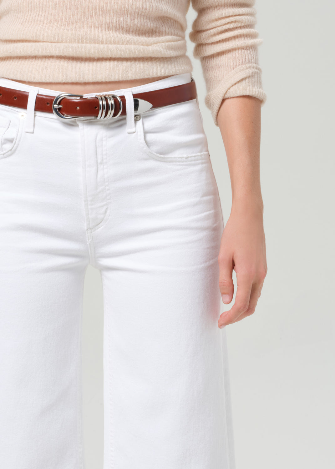 Person wearing a beige sweater, white pants, and a brown belt on a plain background