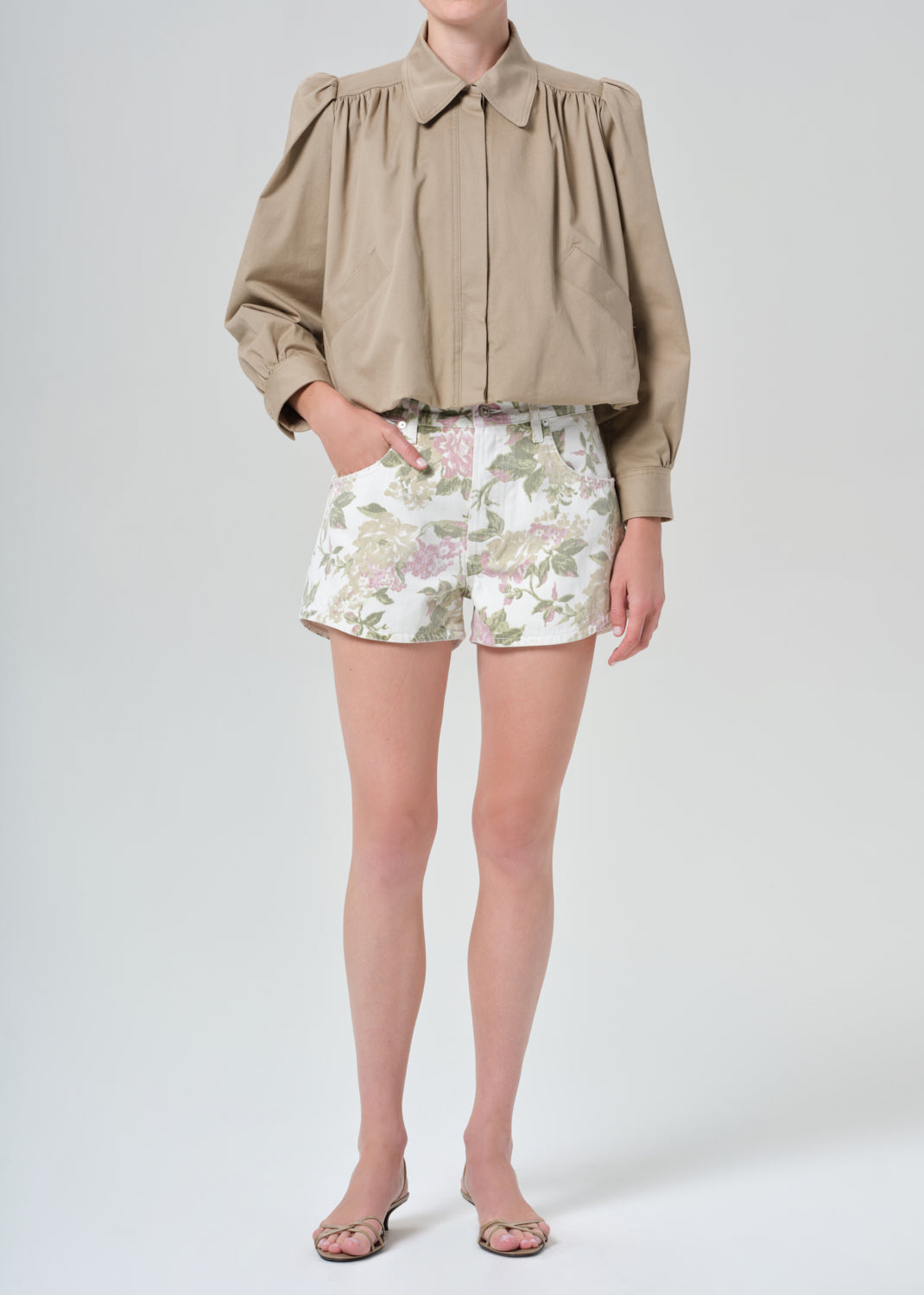 Person wearing a beige jacket and floral  denim shorts on a grey background