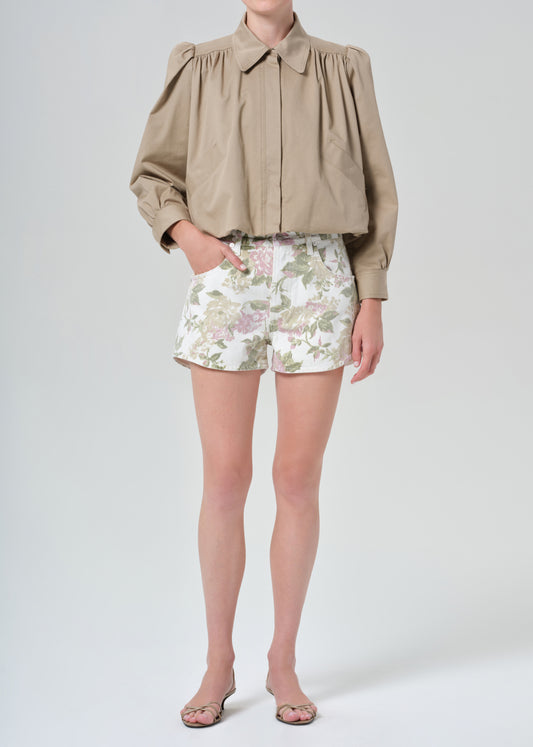 Person wearing a beige jacket and floral  denim shorts on a grey background