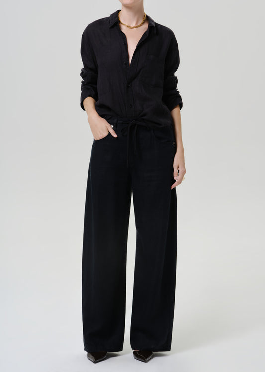 Person wearing black wide leg pants with a black long sleeve button-up blouse and black pointed-toe heels, front full body view on a grey background.