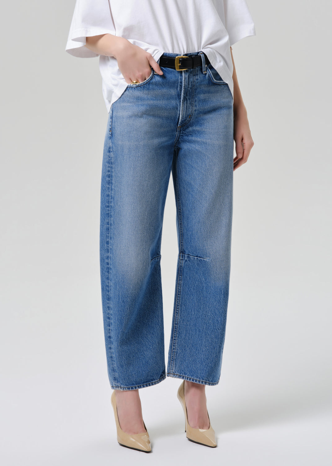 A model wearing a pair of wide-leg cropped blue jeans with a faded wash, styled with a white t-shirt, black belt, and beige heels against a grey background.
