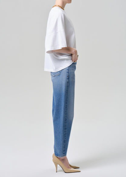 A side-profile studio shot of a model wearing a white short-sleeve top, light-wash blue jeans, and beige high-heeled pumps against a grey background.
