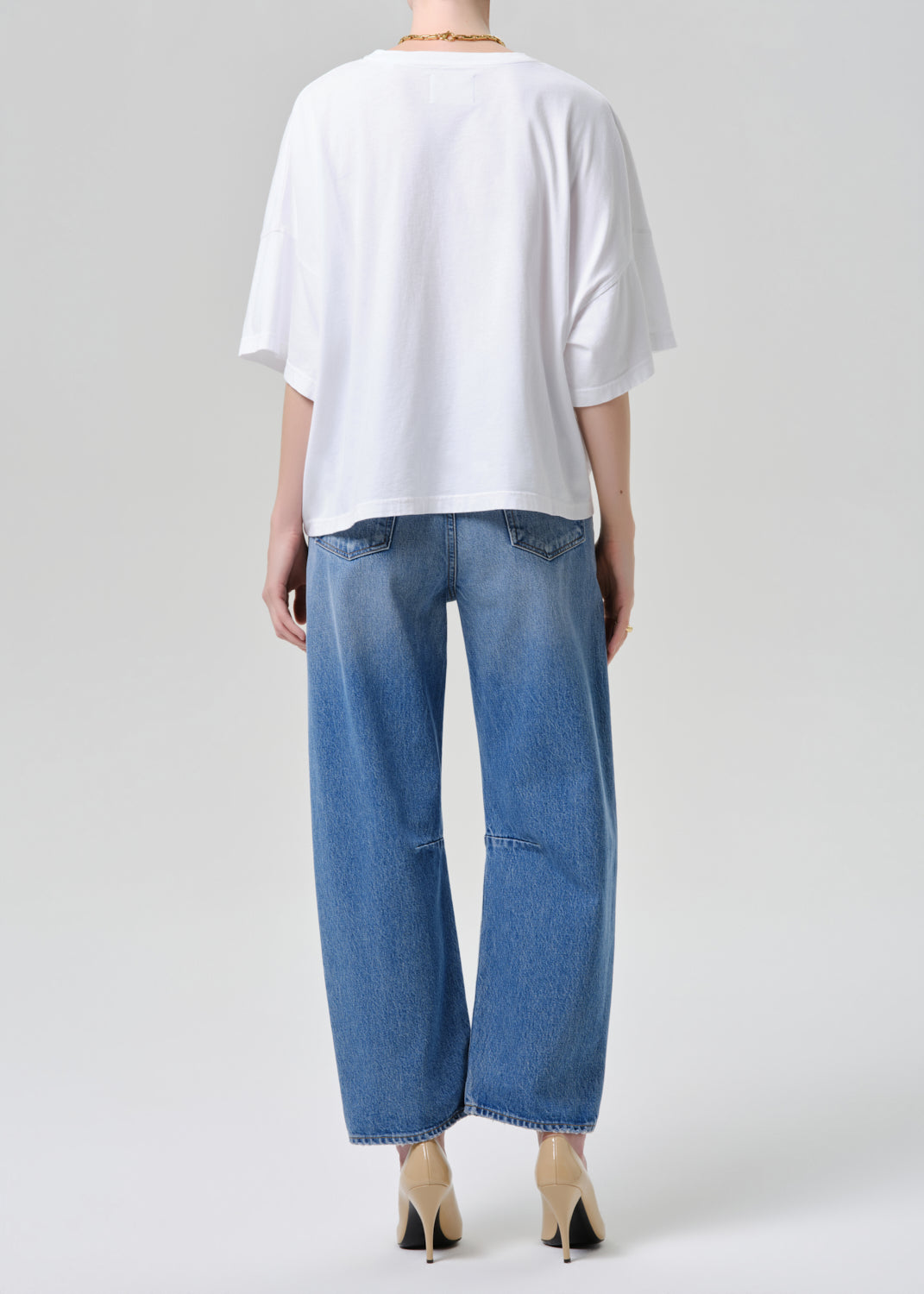 A model shown from the back wearing a white oversized short-sleeve t-shirt, blue wide-leg cropped jeans, and beige pointed-toe heels against a grey background.