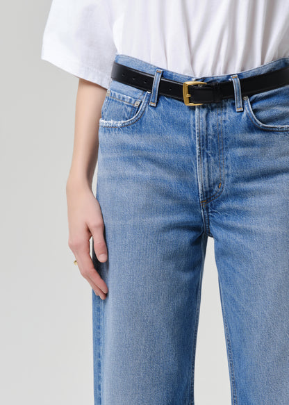 Close-up of a person wearing light blue denim jeans with a black belt and gold buckle, paired with a white top, against a grey background.