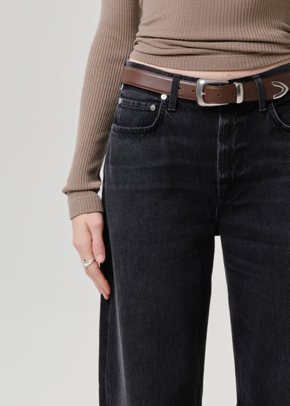Close-up of a person wearing dark washed wide-leg jeans, a brown leather belt with a silver buckle, and a ribbed taupe long-sleeve top against a light grey background.