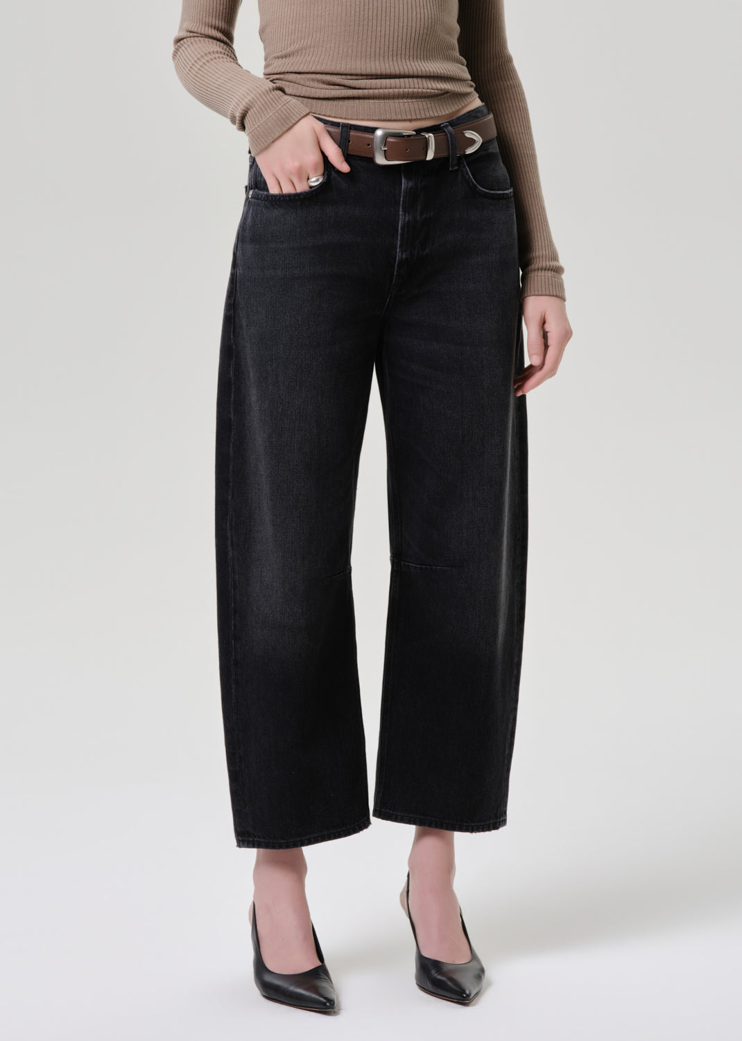 A model wearing black wide-leg cropped jeans with a brown belt, paired with a beige ribbed long-sleeve top and black pointed-toe heels, photographed against a light grey background.