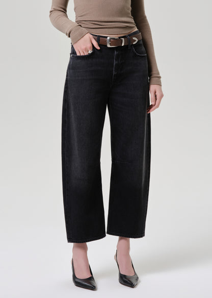 A model wearing black wide-leg cropped jeans with a brown belt, paired with a beige ribbed long-sleeve top and black pointed-toe heels, photographed against a light grey background.