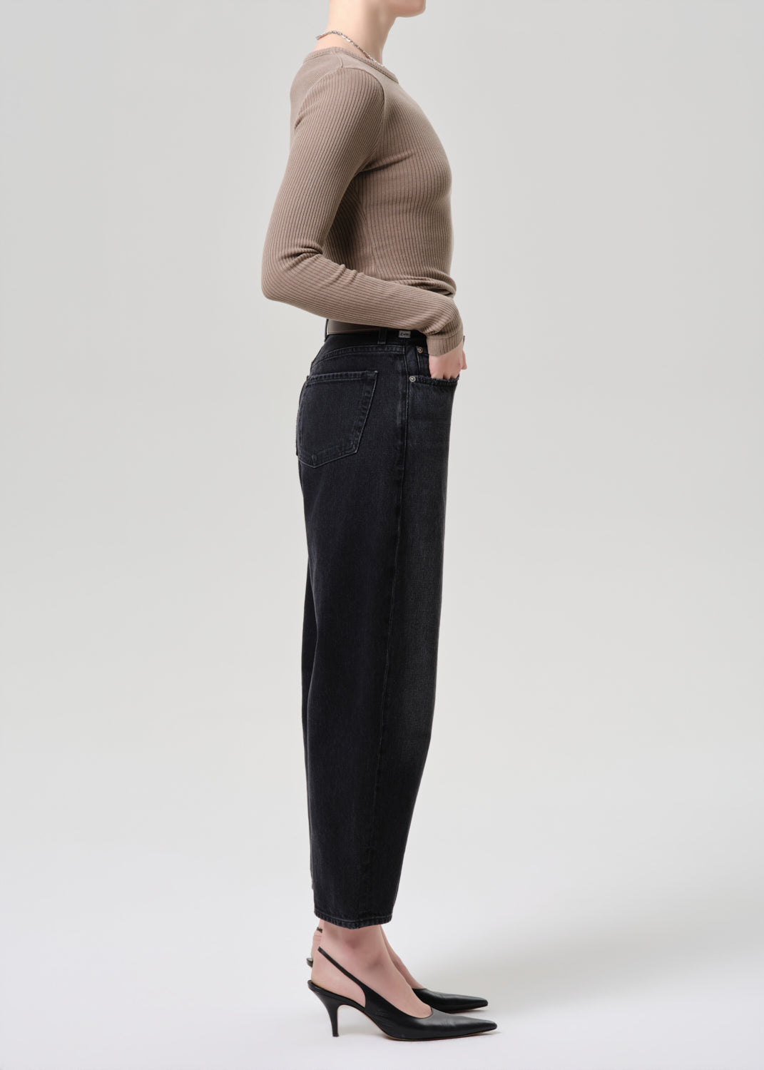 A side-profile studio shot of a model wearing a beige ribbed long-sleeve top, black wide-leg cropped jeans, and black slingback heels against a light grey background.