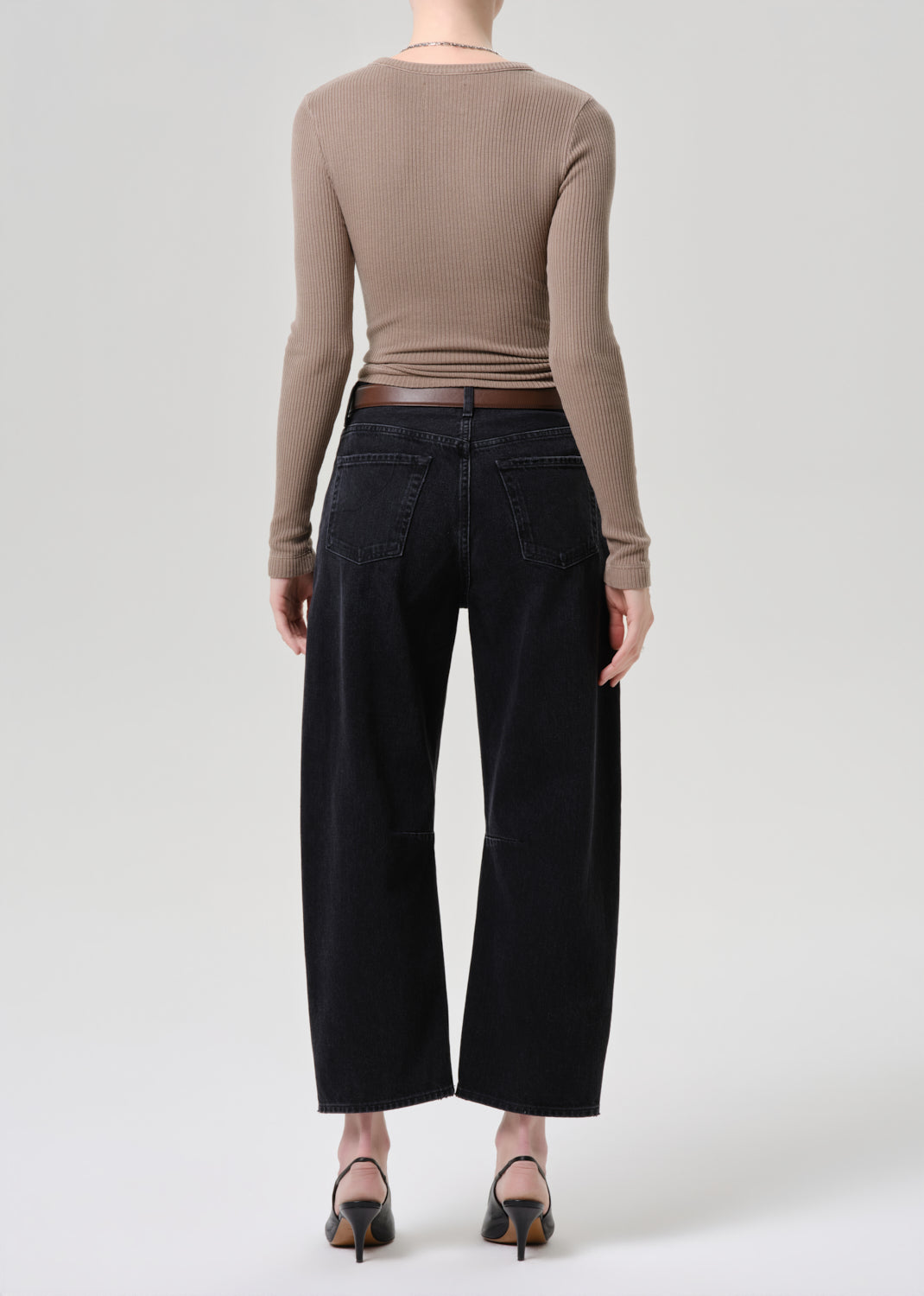 A model shown from the back wearing a beige long-sleeve top, black wide-leg cropped jeans, and black pointed-toe heels against a light grey background.