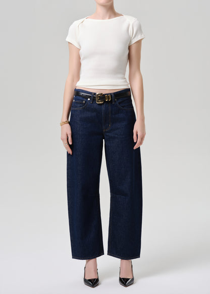A model wearing dark blue wide-leg cropped jeans with a gold-tone belt, paired with a white short-sleeve top and black pointed-toe heels, photographed against a grey background.
