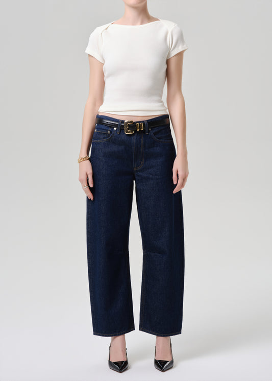 A model wearing dark blue wide-leg cropped jeans with a gold-tone belt, paired with a white short-sleeve top and black pointed-toe heels, photographed against a grey background.