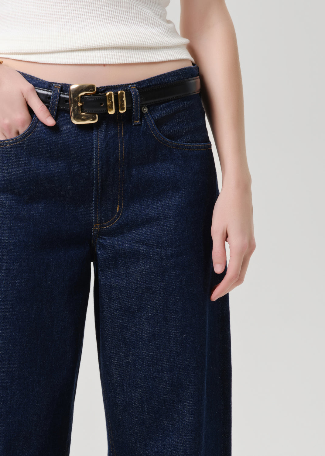 A close-up of a person wearing dark blue denim jeans with a black leather belt featuring a prominent gold buckle, paired with a cream ribbed top against a grey background.